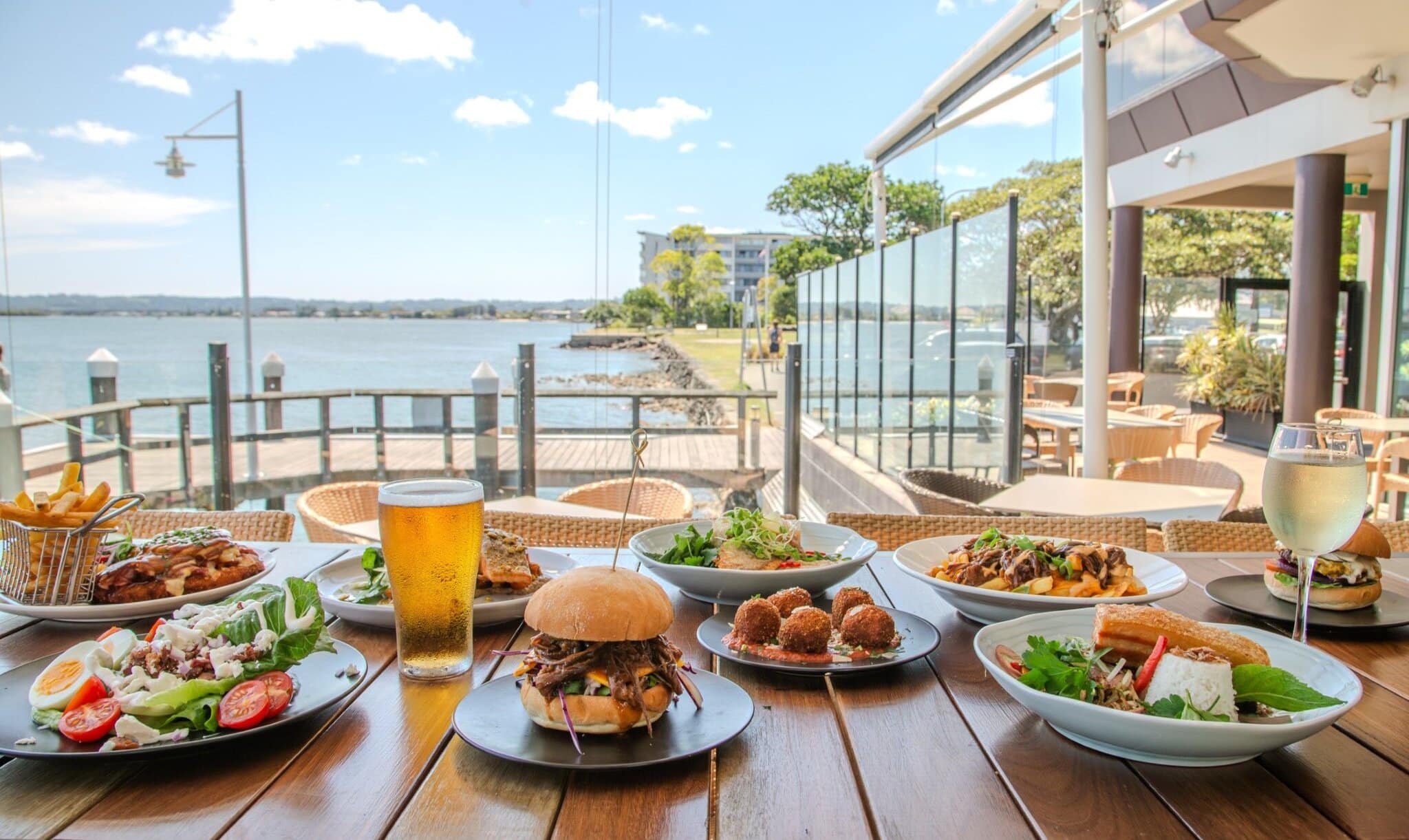Gourmet meals, burgers, salads, and drinks served on a waterfront deck at Ballina RSL, overlooking the Richmond River on a sunny day.
