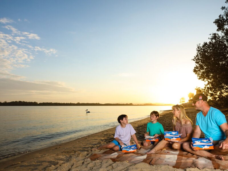 Family enjoying fish and chips picnic on the riverbank at sunset in Ballina, with a pelican gliding across the water.