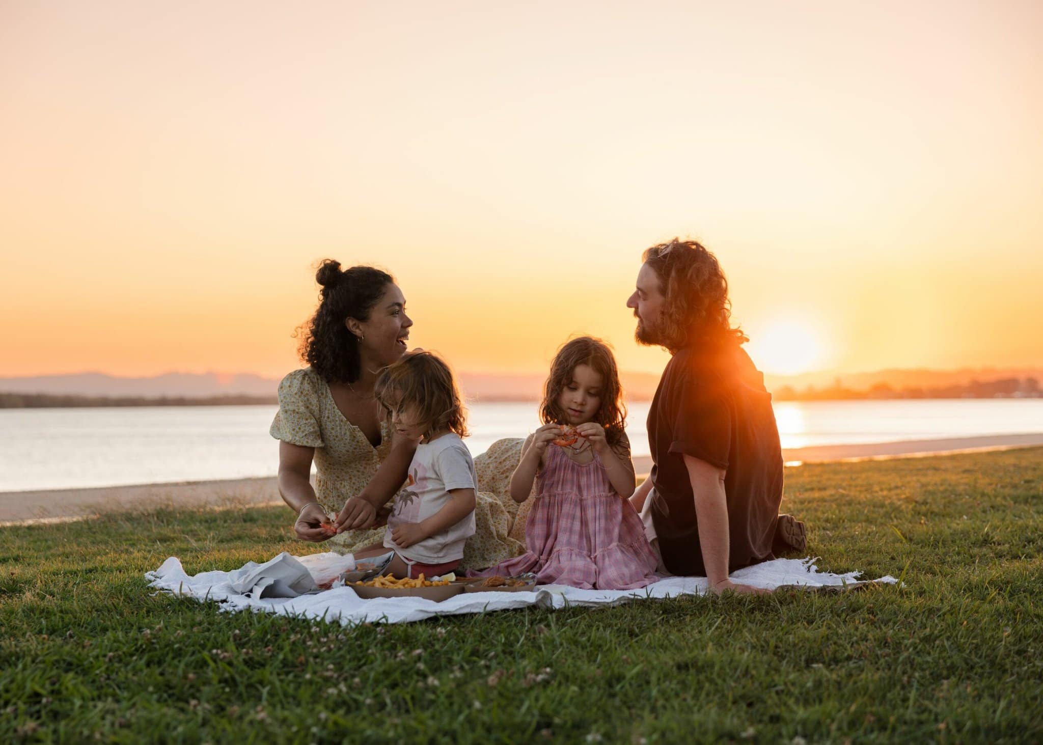 Young family enjoying a picnic on the grass by the Richmond River at sunset in Ballina, with golden skies and water views in the background.