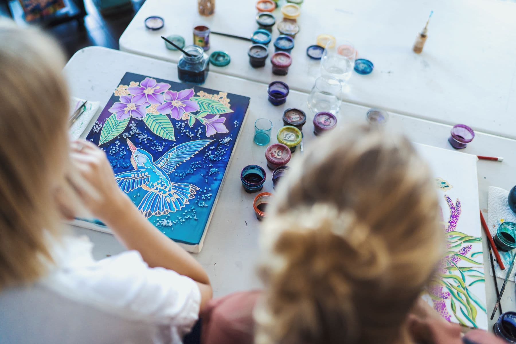 Two participants working on vibrant silk paintings at a Kim Toft workshop, with colourful paint pots and brushes spread across the table.