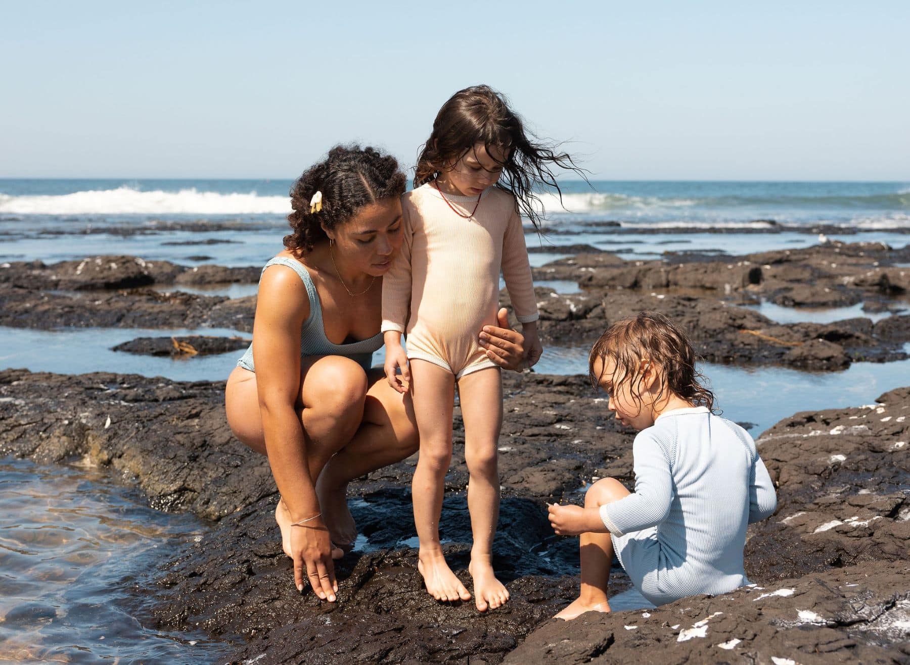 A mother and two children exploring rockpools on a volcanic rock shelf by the ocean in Ballina on a sunny day.