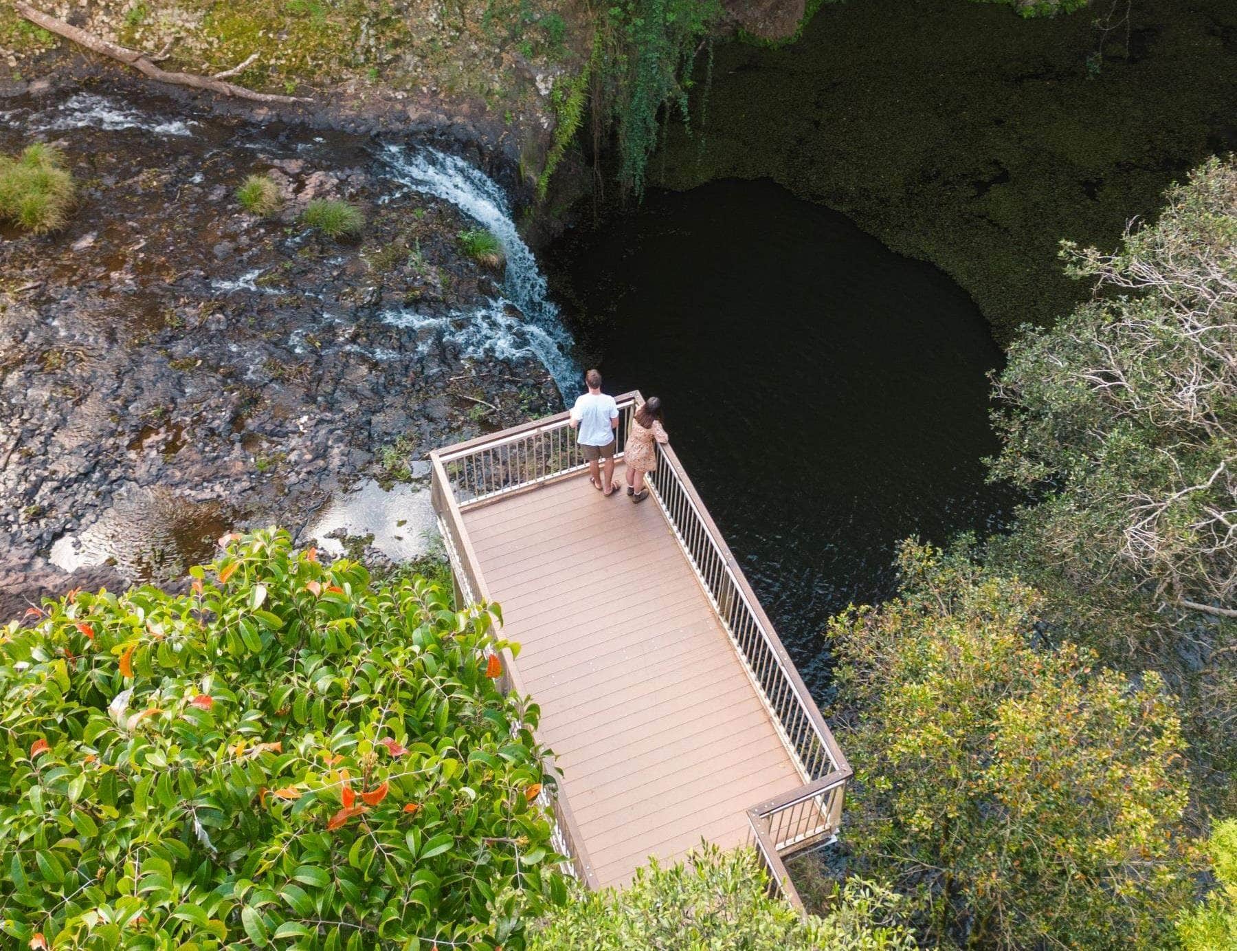 A couple standing on a wooden viewing platform overlooking the lush gorge and plunge pool of Killen Falls in the Ballina Hinterland, surrounded by dense rainforest.