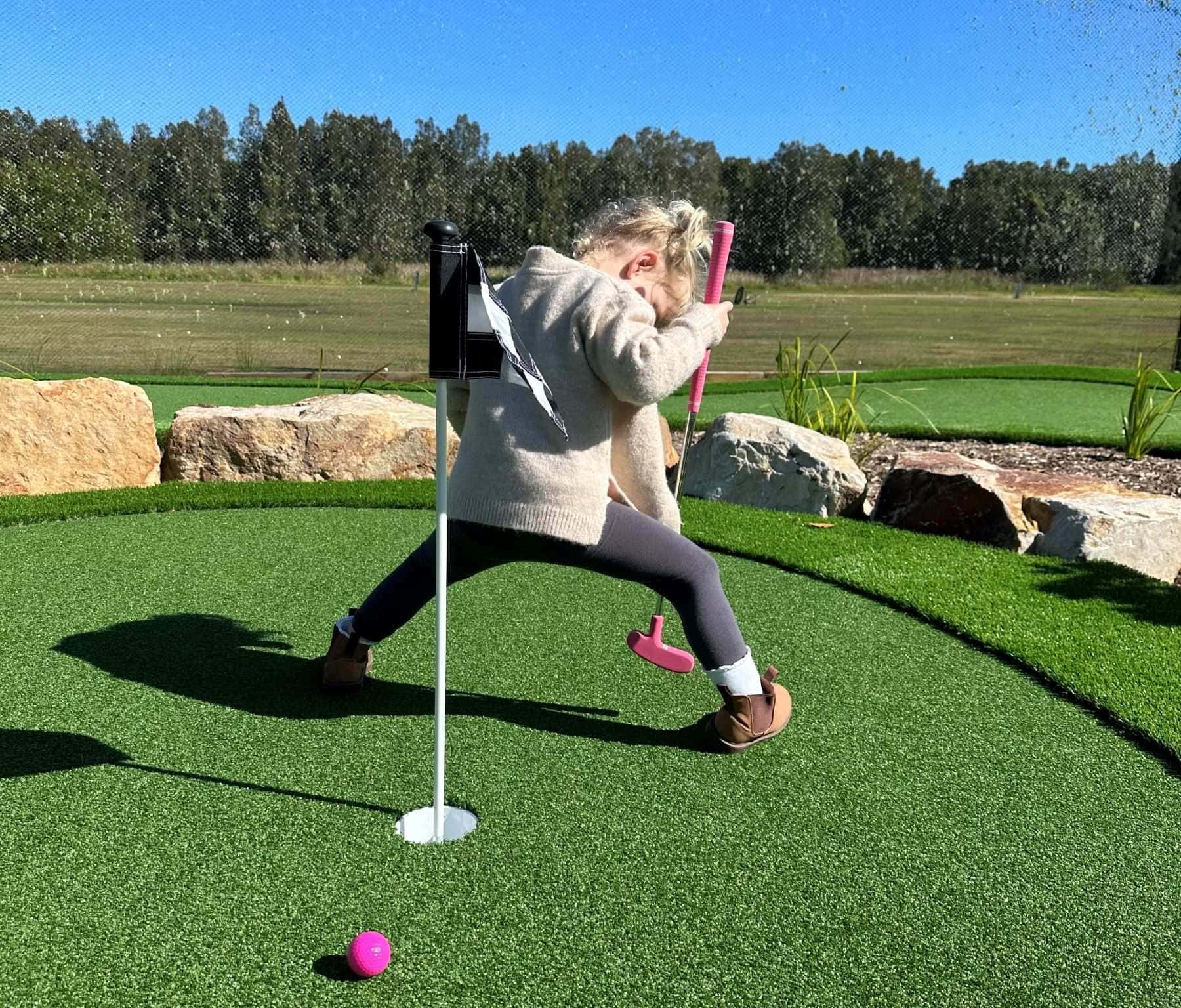 Young child playing mini golf at Swing It Golf Ballina, celebrating a putt on the artificial green with a bright pink golf ball and club.