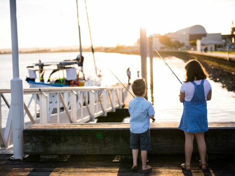 Two children fishing off a wooden jetty on the Richmond River at sunset, with boats moored nearby and a waterfront path in the background.