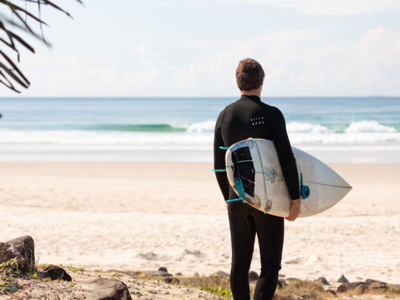 Surfer in wetsuit holding surfboard and looking out at waves on a sandy beach in Ballina, NSW