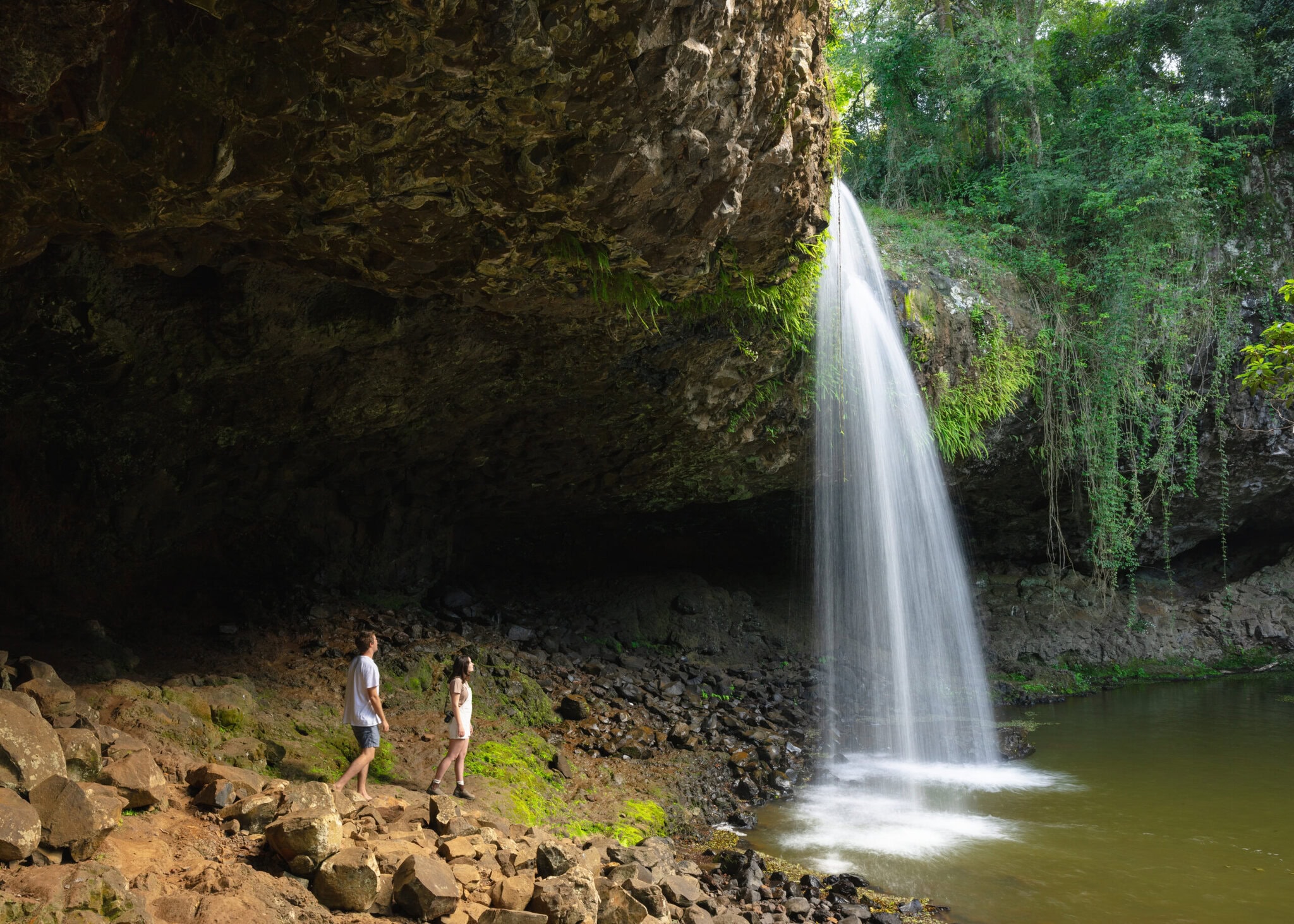 Two people standing beneath the dramatic overhang of Killen Falls in the Ballina hinterland, surrounded by rainforest and a flowing waterfall.