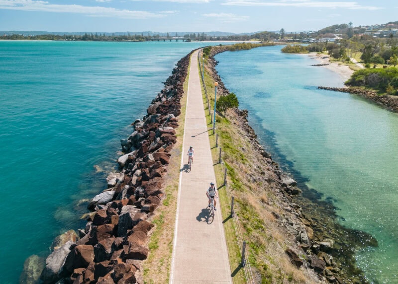 Two cyclists ride along Ballina’s North Wall shared path between the Richmond River and the Pacific Ocean on a sunny day.