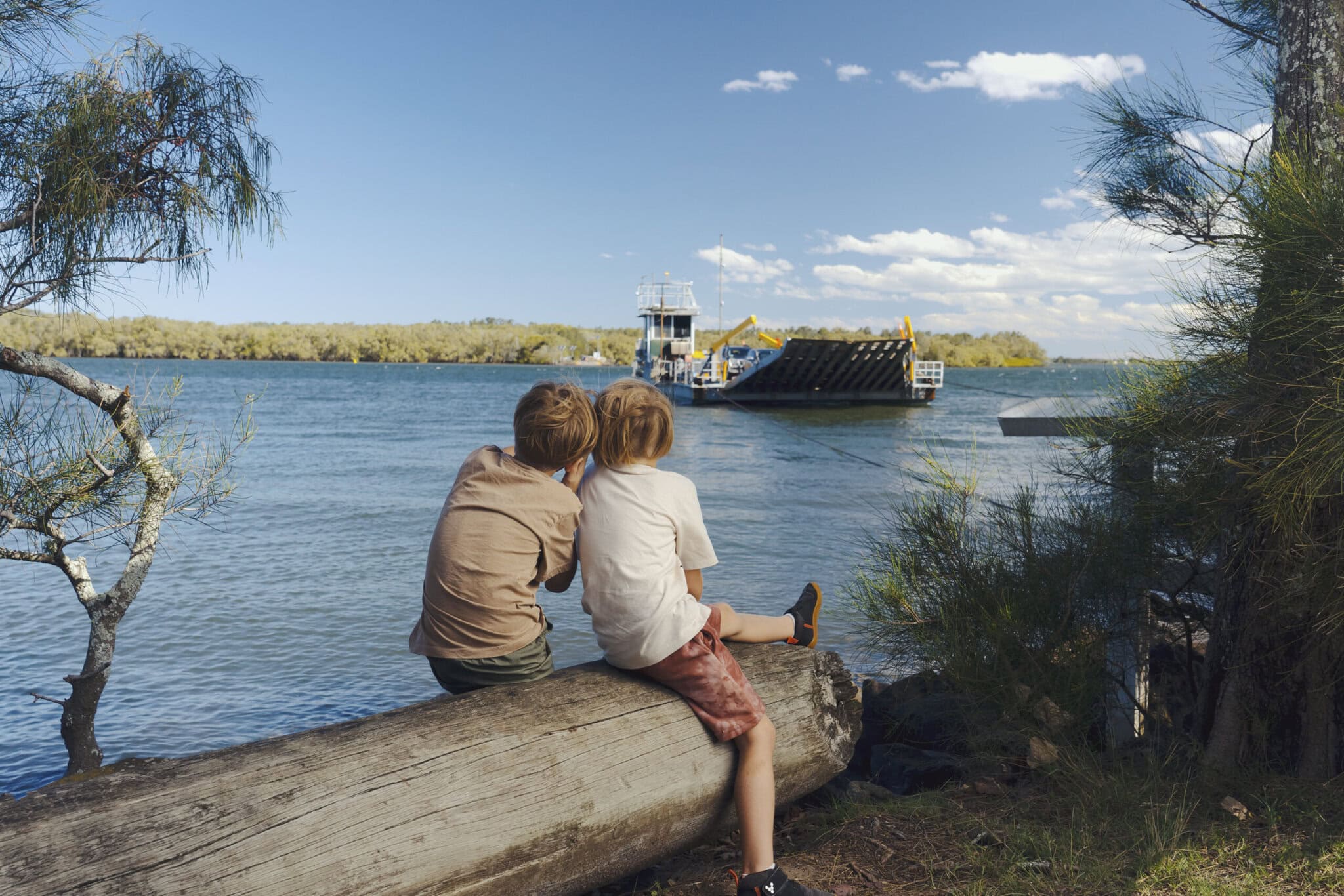 Two children sit on a log by the riverbank, watching the Burns Point Ferry cross the Richmond River in Ballina on a sunny day.