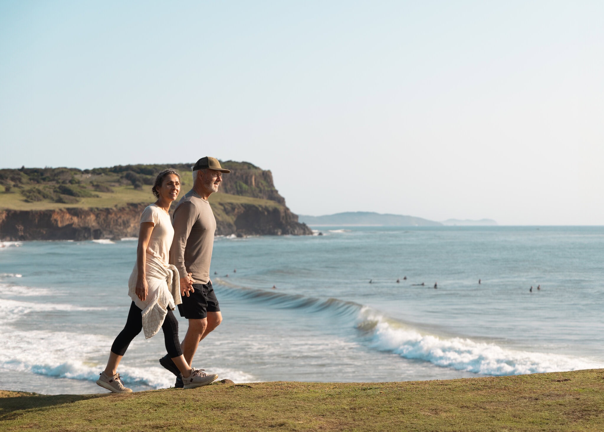 Mature couple walking along the scenic coastal trail at Boulders Beach in Ballina, with ocean waves and surfers in the background.