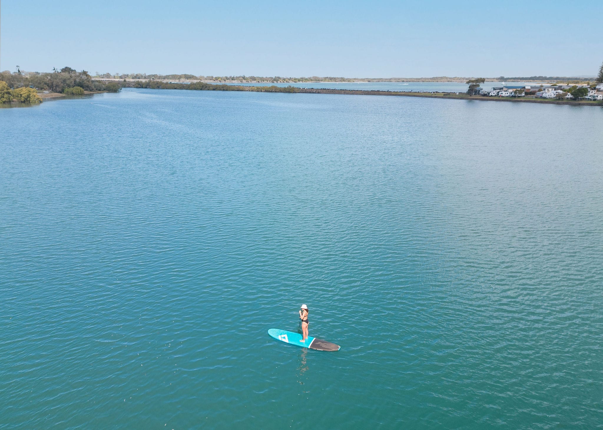 A woman on a stand-up paddleboard glides across the calm, turquoise waters of Shaws Bay in Ballina under a clear blue sky.