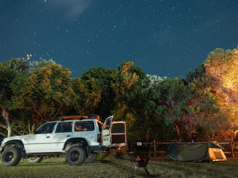 4WD vehicle and swag tent set up under a starlit sky at Flat Rock Tent Park in Ballina, surrounded by trees and a glowing campfire.