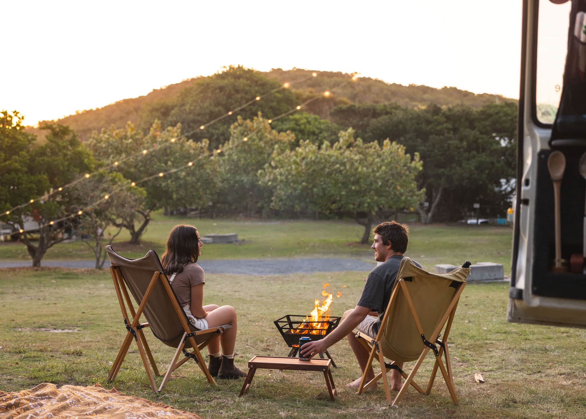 Couple sitting by a campfire under string lights at Flat Rock Tent Park in Ballina, enjoying a peaceful sunset surrounded by trees and hills.