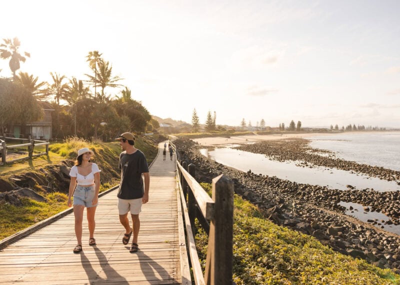 Couple walking along the coastal boardwalk at Lennox Head at sunset, with ocean views, rocky shoreline, and palm trees in the background.