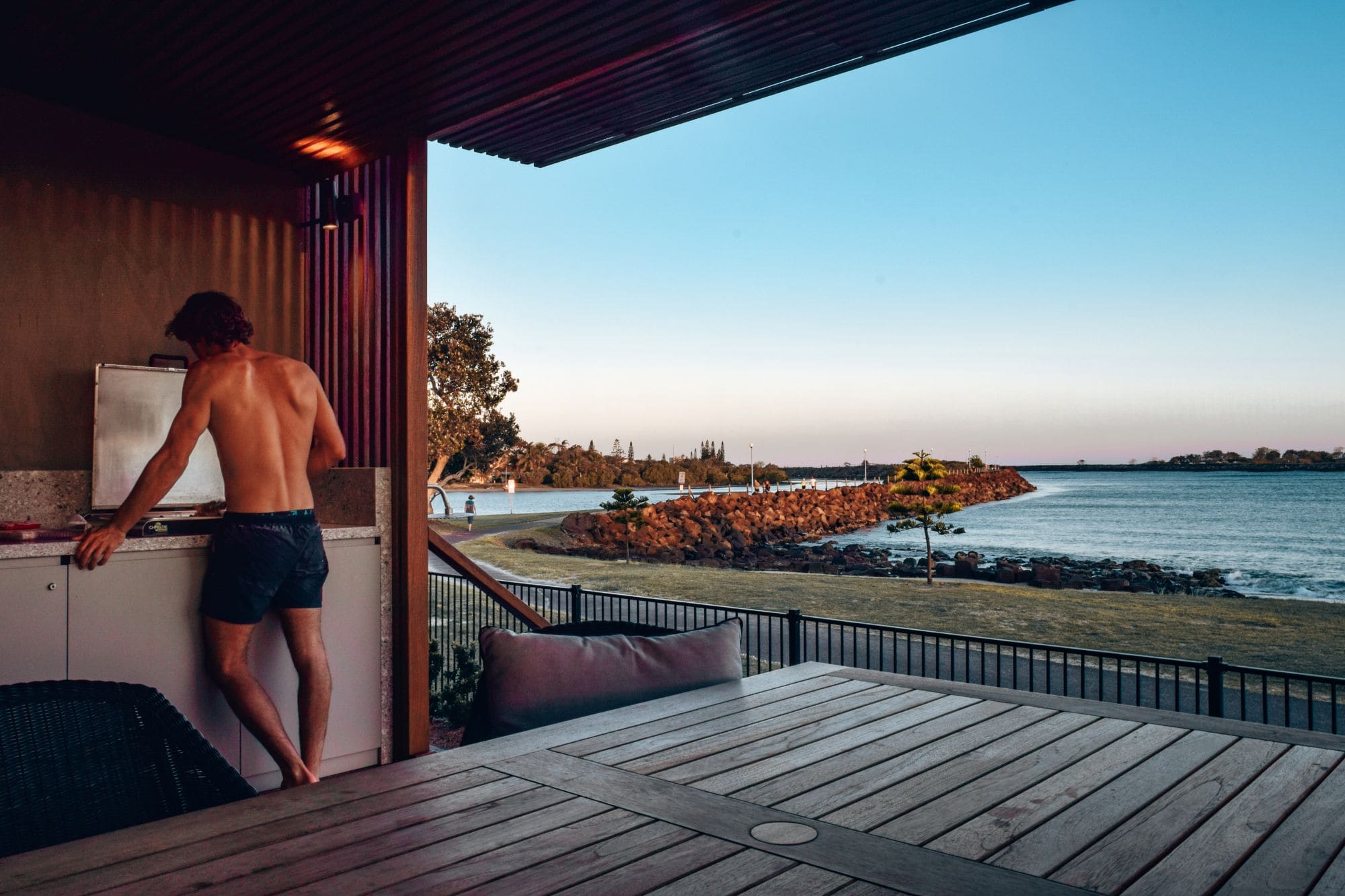 Man cooking at a BBQ in a waterfront cabin at Reflections Holiday Park Shaws Bay, Ballina, with views over the Richmond River at sunset.