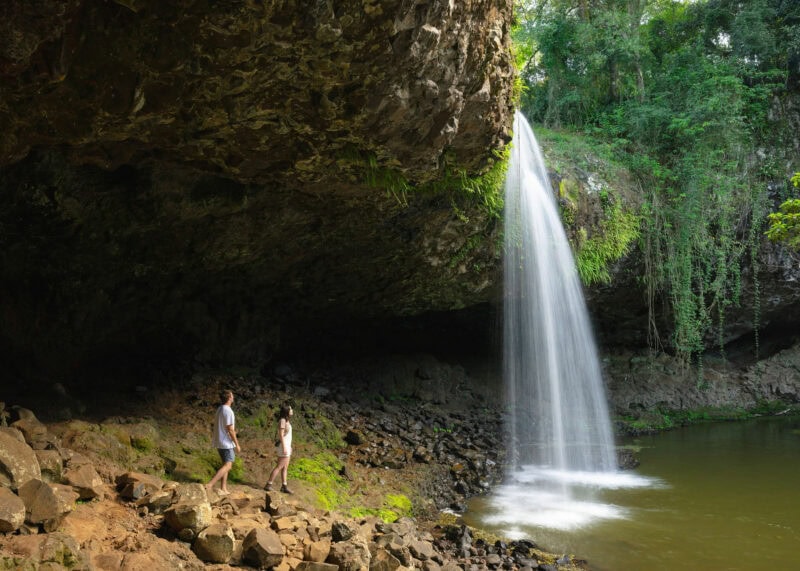 Two people exploring the base of Killen Falls near Ballina, Northern NSW, with water cascading from a cave ledge into a freshwater pool