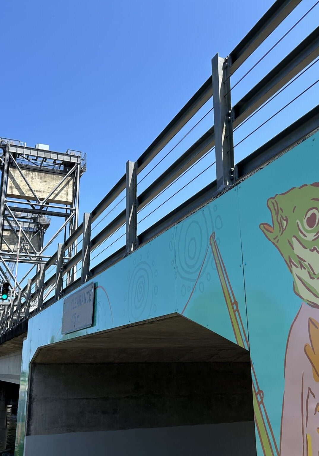 Public mural of a fish-headed figure in a suit beneath the Wardell lift bridge, with bright blue sky and river in view.