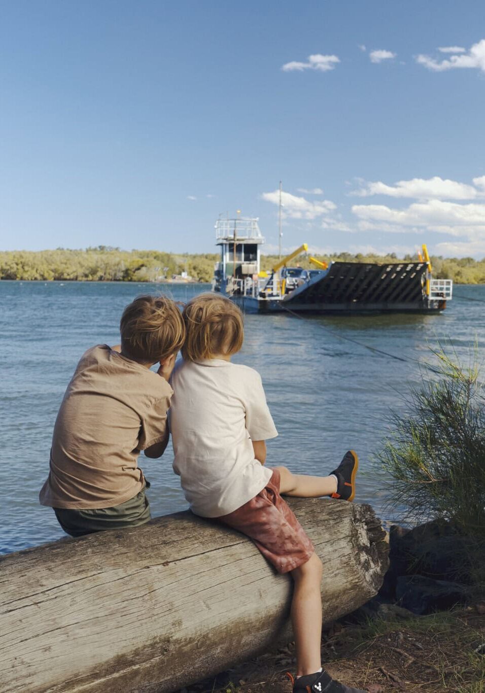 Two children sit on a log by the riverbank, watching the Burns Point Ferry cross the Richmond River in Ballina on a sunny day.