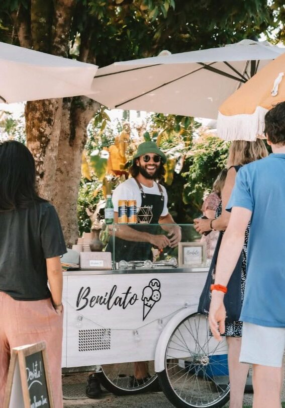 Smiling vendor serving gelato from a Benilato cart under umbrellas at the Newrybar Twilight Markets, surrounded by customers.