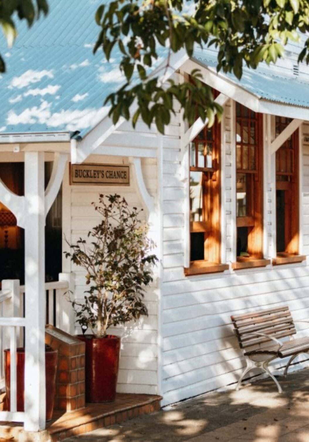 Exterior of Buckley’s Chance café in Alstonville, featuring white weatherboard walls, timber windows, and shaded bench seating under leafy trees.