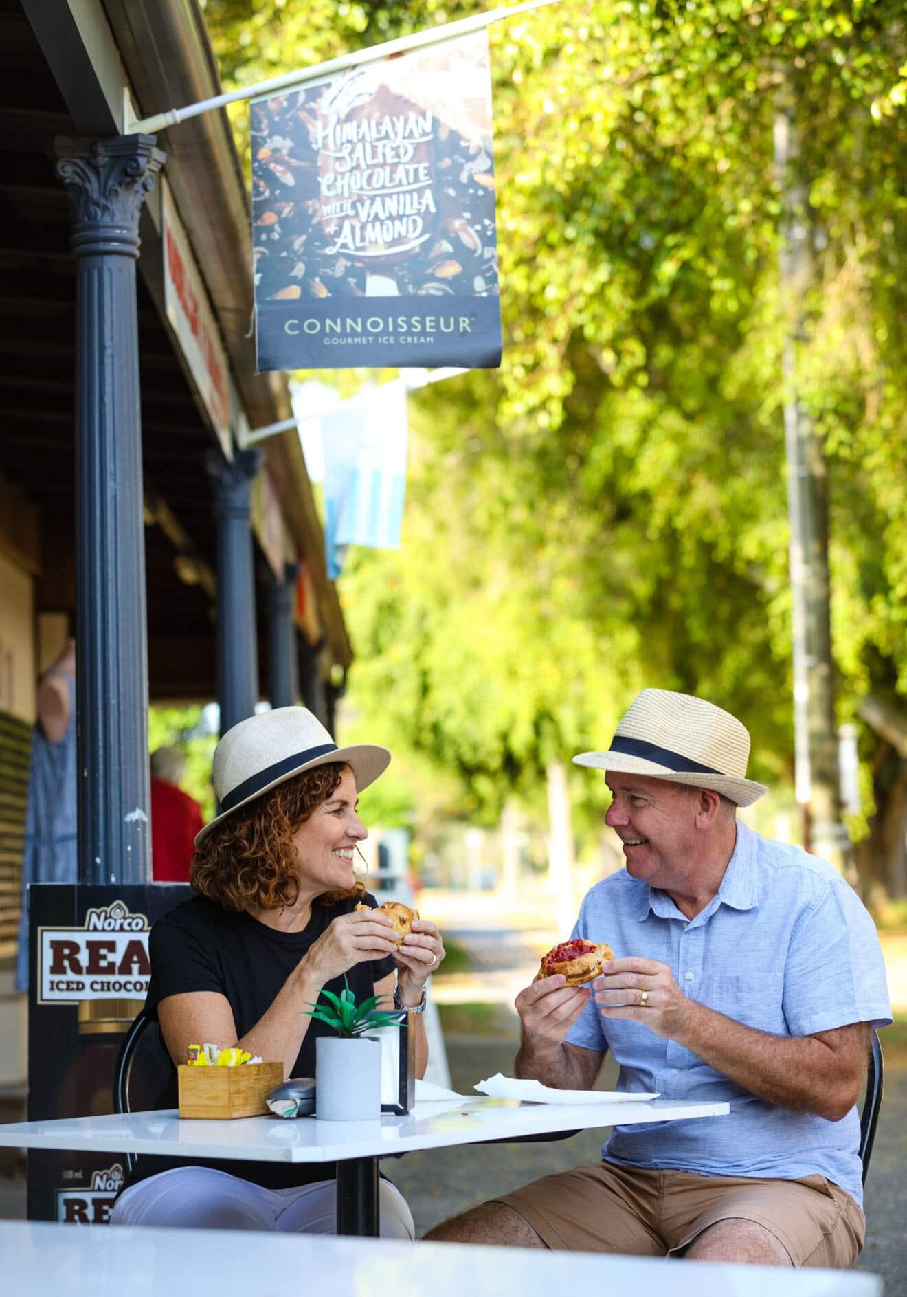 Couple smiling and enjoying freshly baked pies at an outdoor table in Wardell, surrounded by leafy streets and heritage shopfronts.