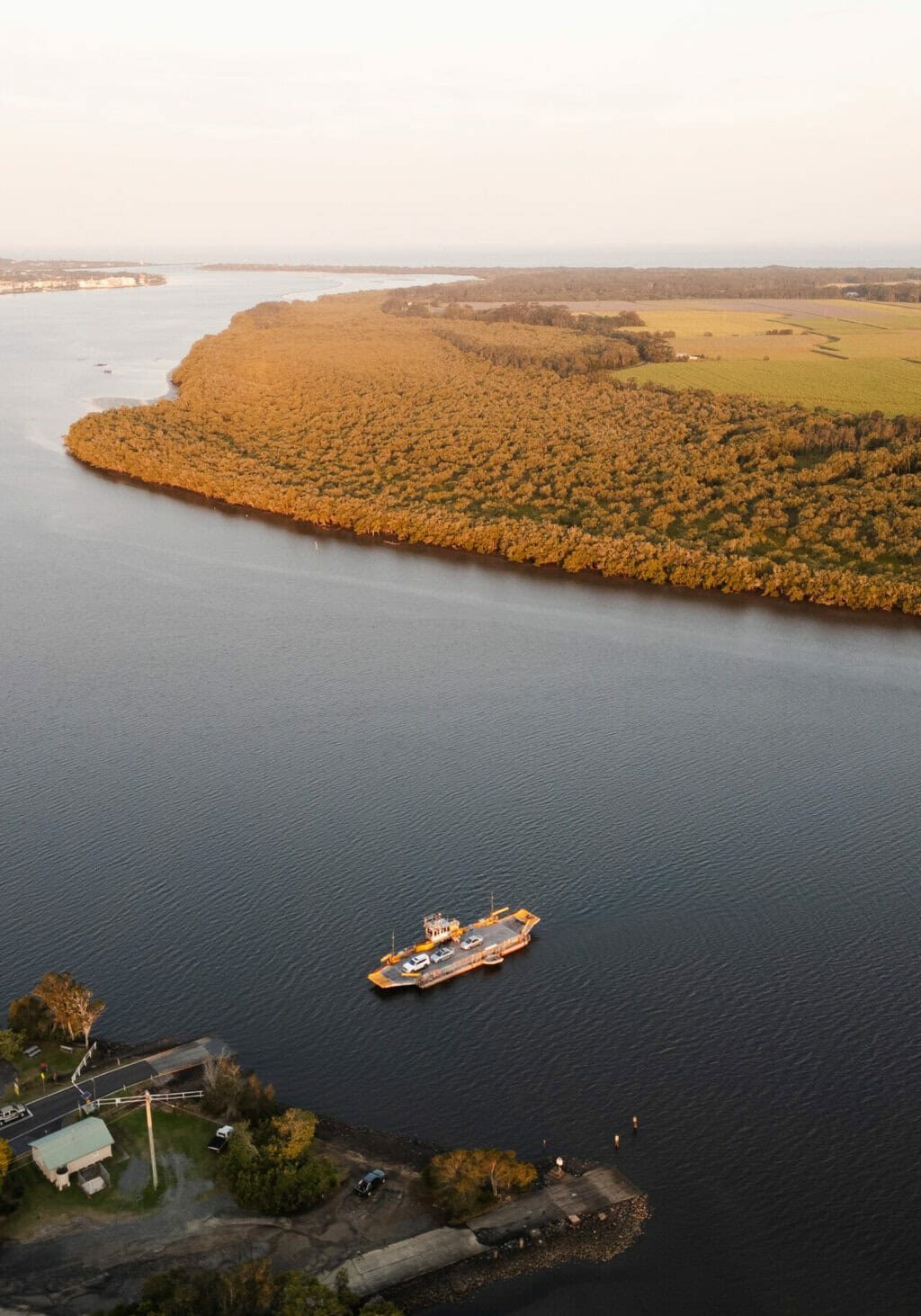 Aerial view of the Burns Point Ferry transporting vehicles across the Richmond River between Ballina and South Ballina, with mangroves and farmland in the background.