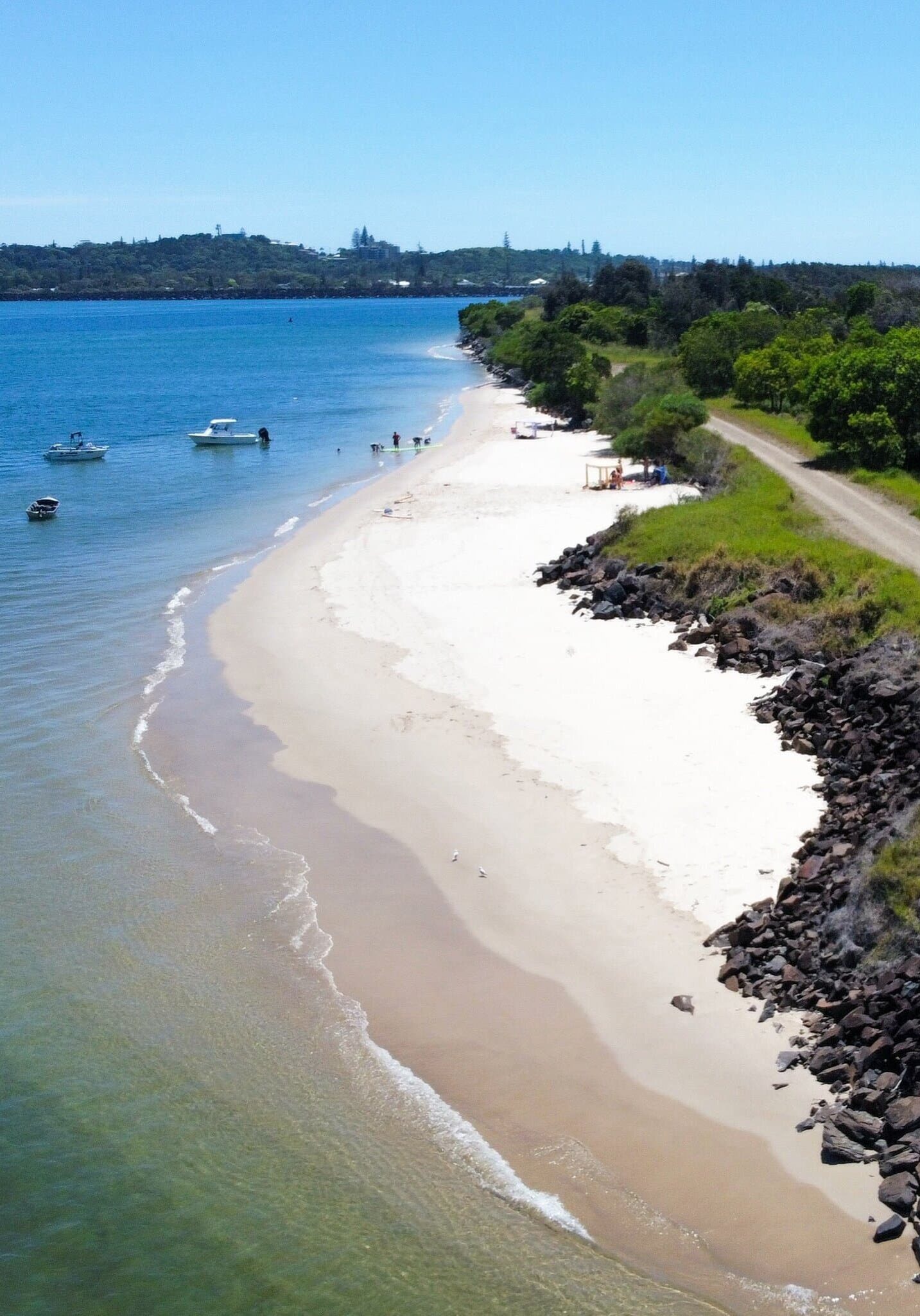 Aerial view of Mobbs Bay in South Ballina, featuring calm turquoise waters, anchored boats, a sandy shoreline, and a coastal track bordered by native bushland.