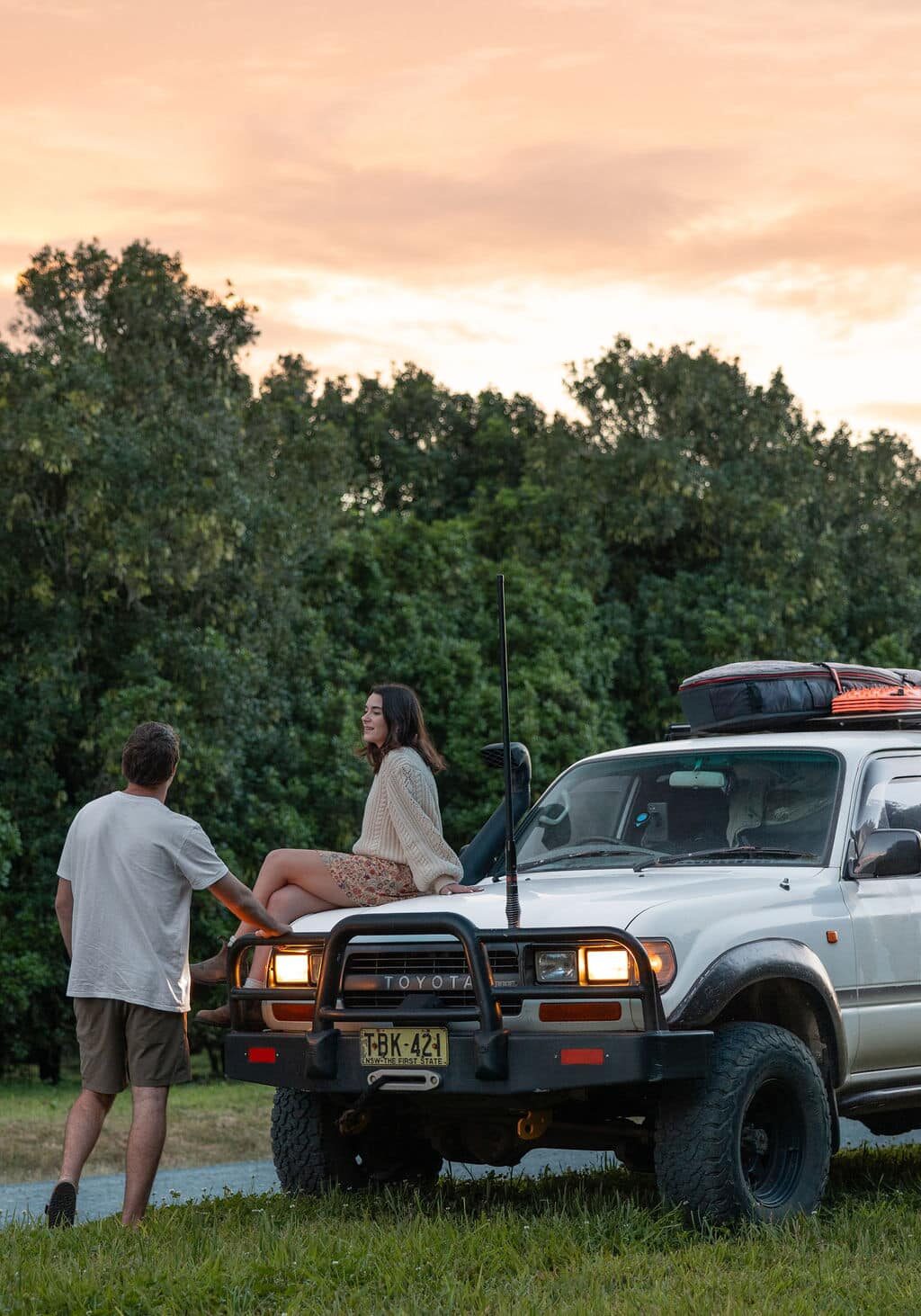 Couple enjoying a quiet moment by their 4WD in the lush hinterland near Ballina, with trees in the background at sunset.